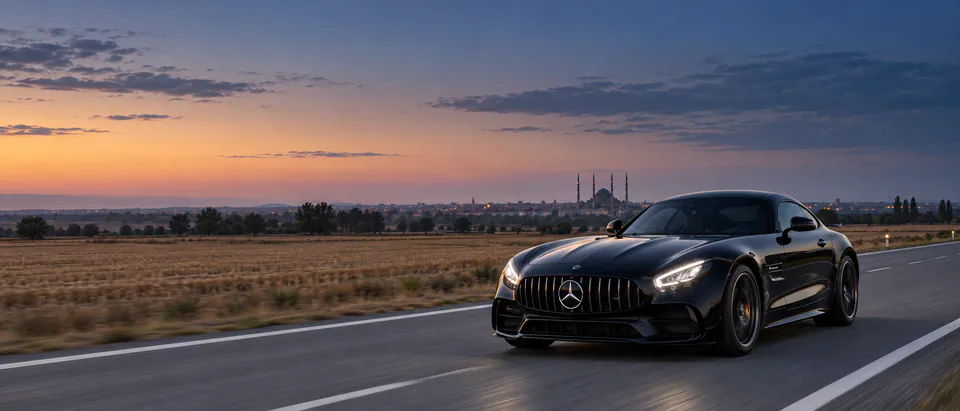 Ova yolunda farları yanan siyah Mercedes-AMG GT; ufukta büyük kubbeli, dört minareli bir caminin silueti