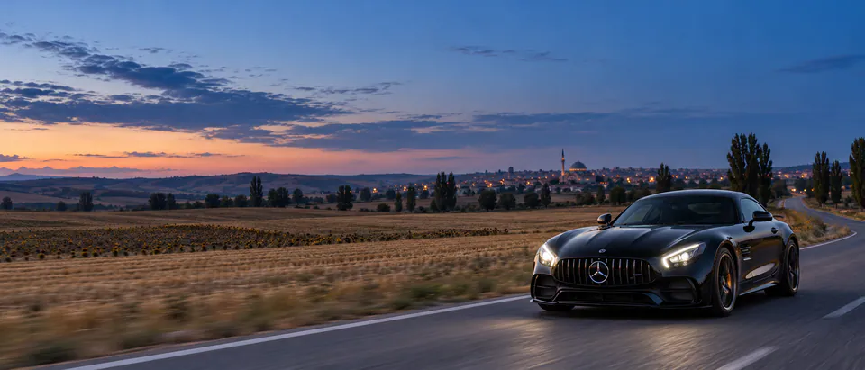Hasat edilmiş tarlalar arasındaki yolda farları yanan siyah Mercedes-AMG GT; ufukta kubbeli, tek minareli bir cami ve kasaba ışıkları